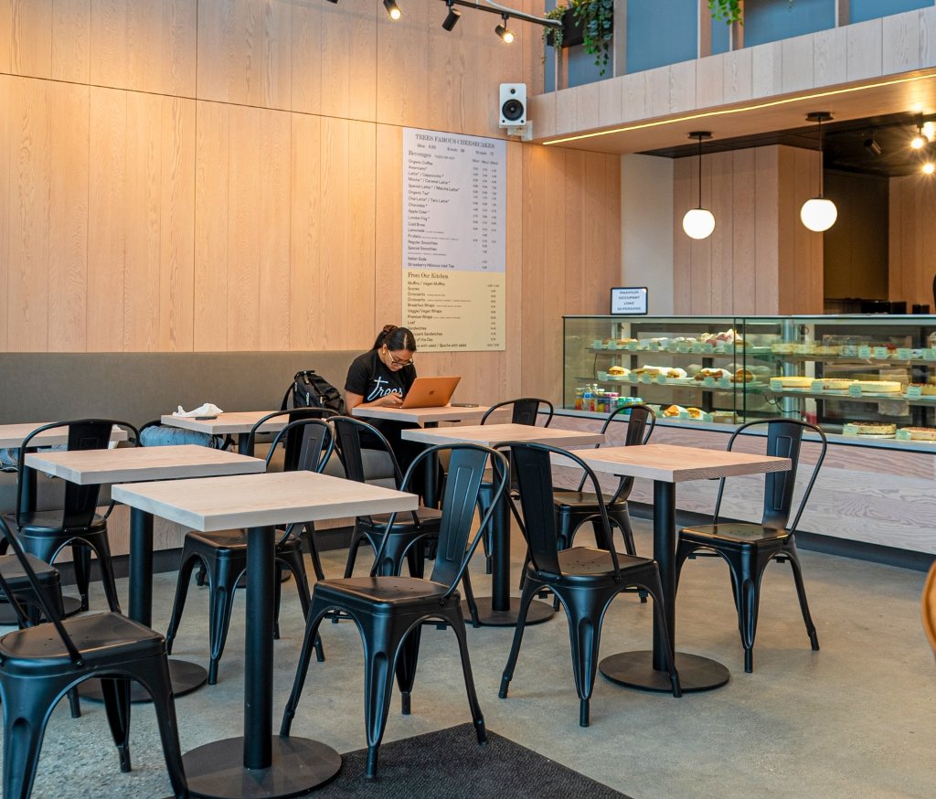 Person typing on a laptop at a table in a modern cafe with wooden walls and a pastry display case in the background.