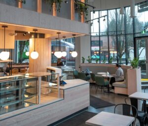 Cafe interior with wooden counters, globe pendant lights, and a person at a booth using a laptop near large windows.