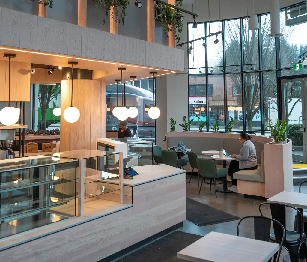Cafe interior with wooden counters, globe pendant lights, and a person at a booth using a laptop near large windows.