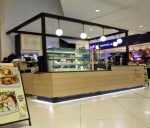 Cafe counter with glass pastry display under a black metal frame and hanging globe lights in a mall food court